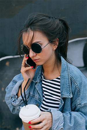 Schmerzen im Daumen: junge Frau mit Zopf, Kaffee to go und Sonnenbrille telefoniert mit dem Smartphone. Bild: Pexels/Katie E